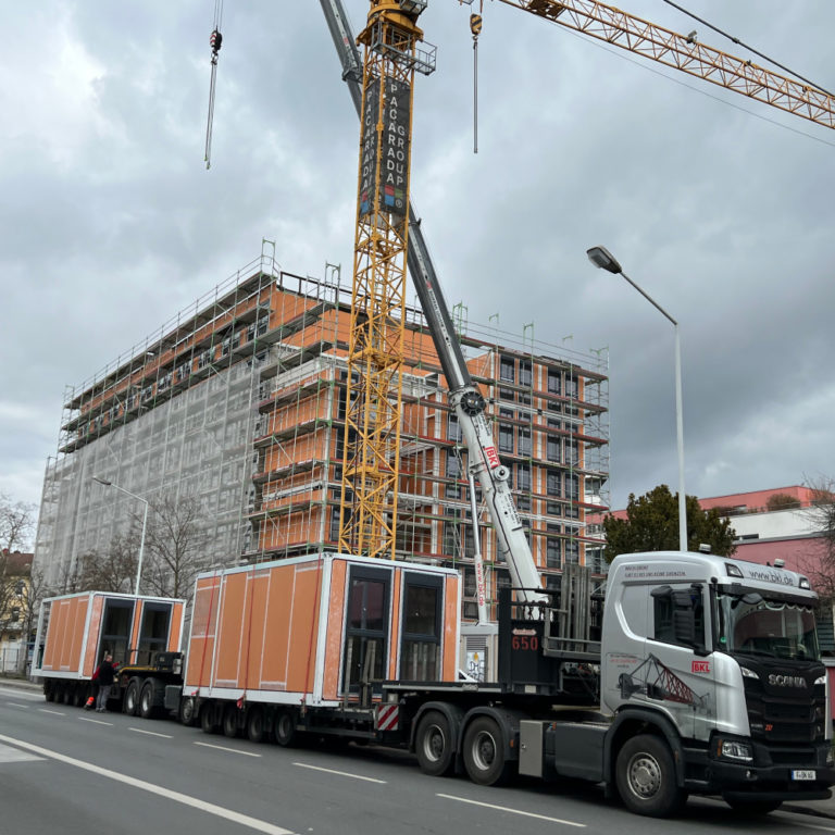 Modulbau in Darmstadt: Module auf dem Tieflader, dahinter Kran und eingerüstetes Gebäude